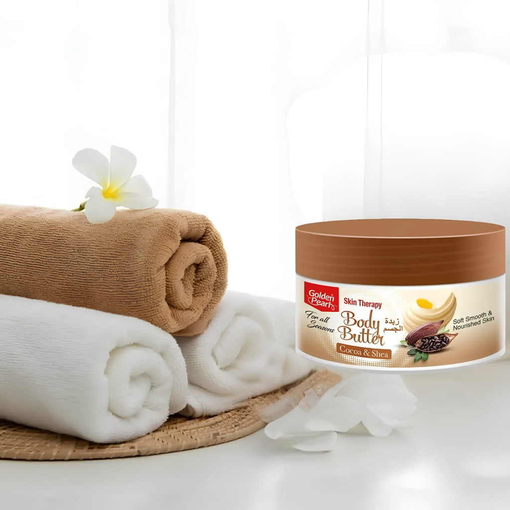 A jar of Golden Leaf Skin Therapy Body Butter sits on a white surface beside rolled towels and a white flower. The product container is brown with a cream label featuring product imagery.