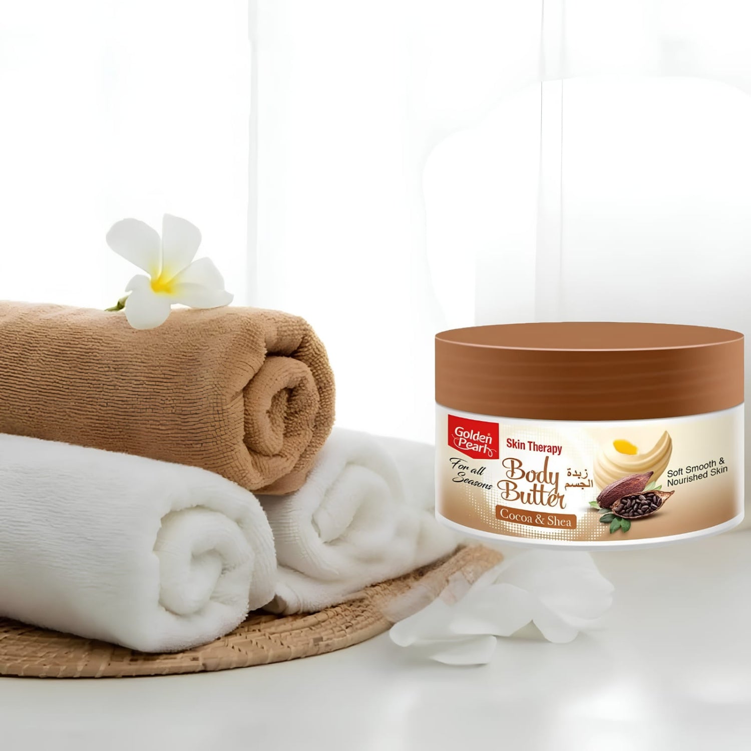 A jar of Golden Leaf Skin Therapy Body Butter sits on a white surface beside rolled towels and a white flower. The product container is brown with a cream label featuring product imagery.