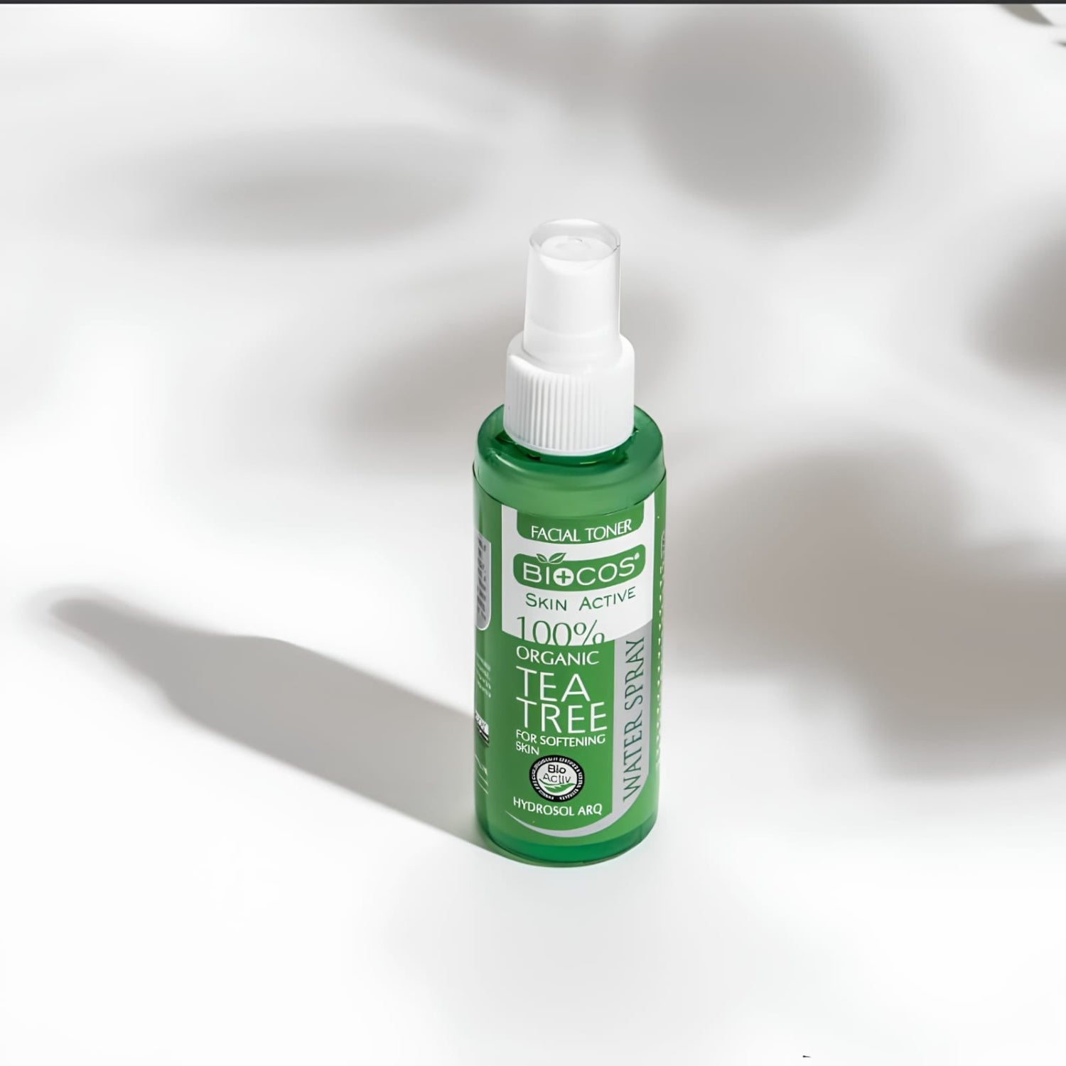 A green bottle of Biotics Skin Active Tea Tree Facial Toner with a white spray cap, set against a white background with soft, artistic shadows.